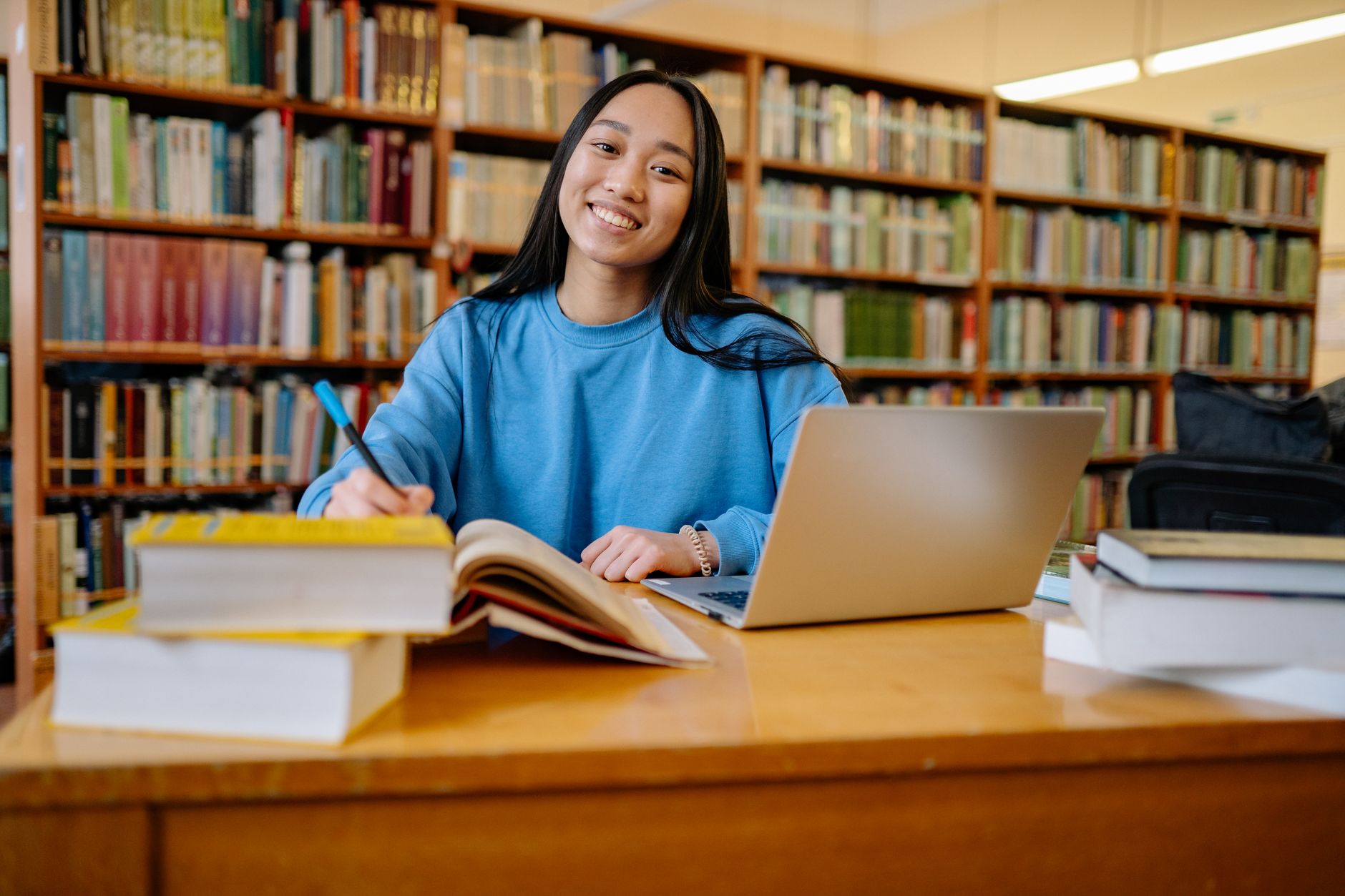 Student writing thesis notes at a library desk with a laptop and open book